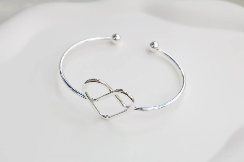 Silver heart knot open bangle bracelet with polished metal finish, double heart charm design and round ball ends for wholesale jewelry buyers